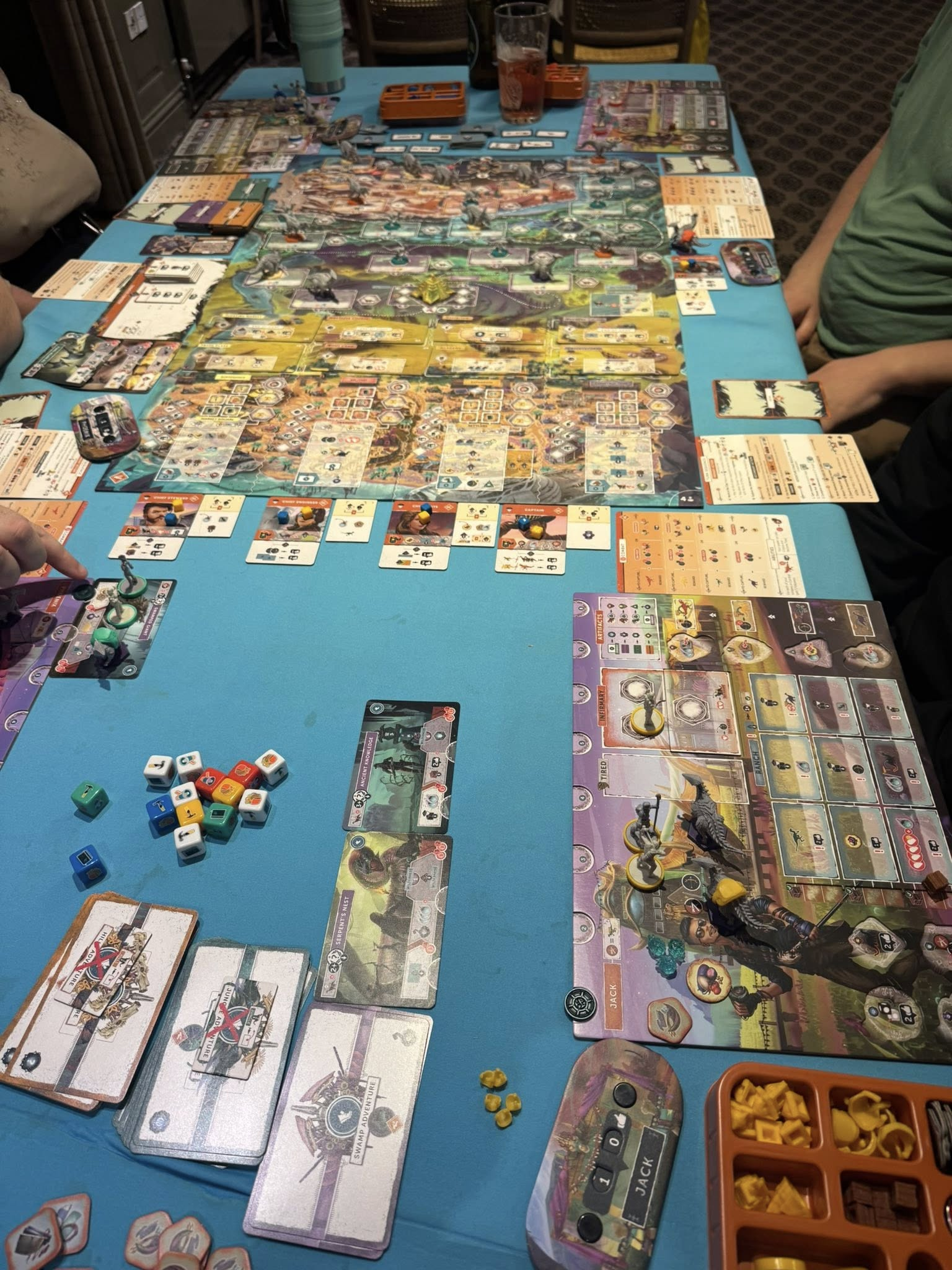 View from the end of a very full table, in the foreground a group of coloured dice with icons on, a player board with some dinosaur and ranger figures, in the background stacks of cards, a large board that shows four columns of city dice-action spots, and a map of wilderness exploration trained-dino spots