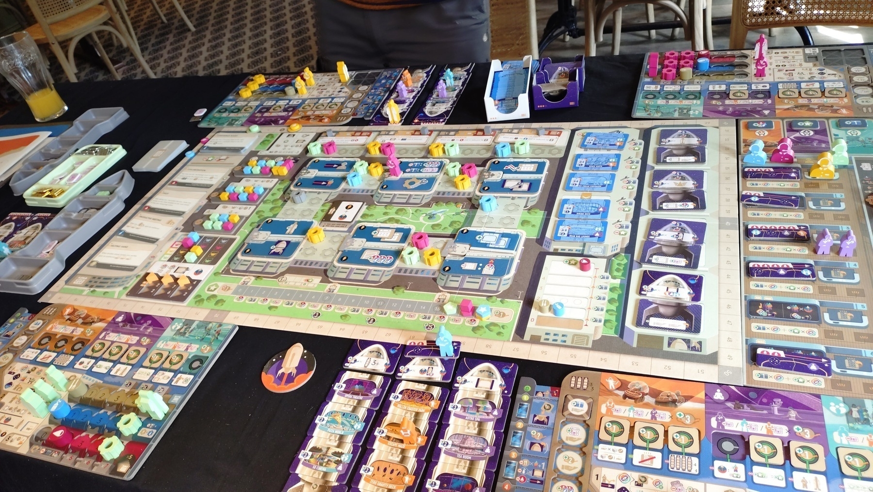 End stage of the game "Galactic Cruise", a large board with pastel coloured design, blue, yellow, green and pink player meeples occupy worker spots for collecting and building spaceship parts, and planning space criuises. Orange, Blue and Purple "guest" meeples wait at the edge of the board. Each player has a sub board tracking their progress and several assembled space ships