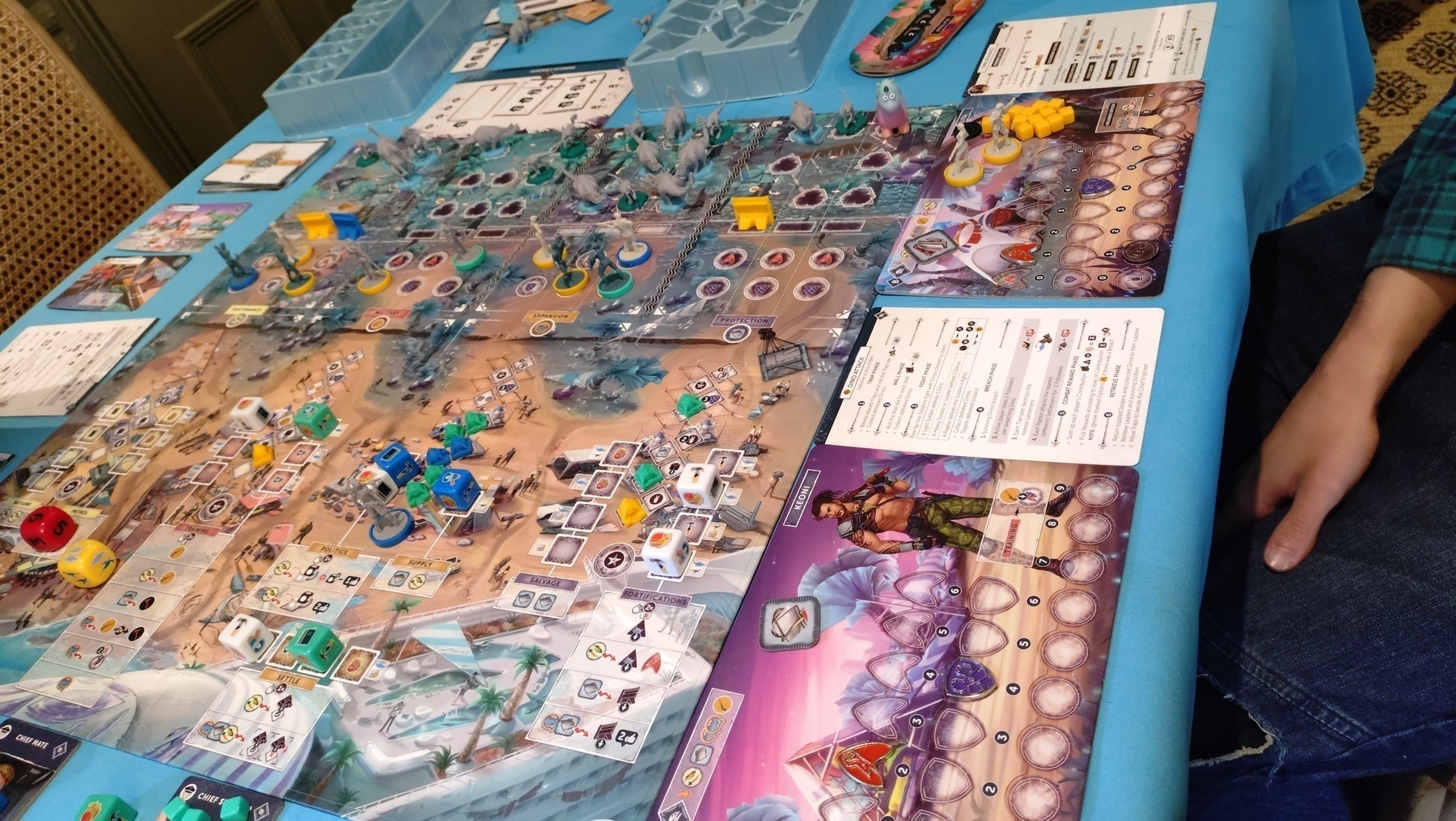 Perseverance, Castaway chronicles, Episode one mid-game. The board shows a beach with various worker-placement (dice) locations which can be used to gather resources, send out soldies on patrol, train more soldiers. After the beach there are defensive positions for soldiers and walls, and attacking dinosaur models