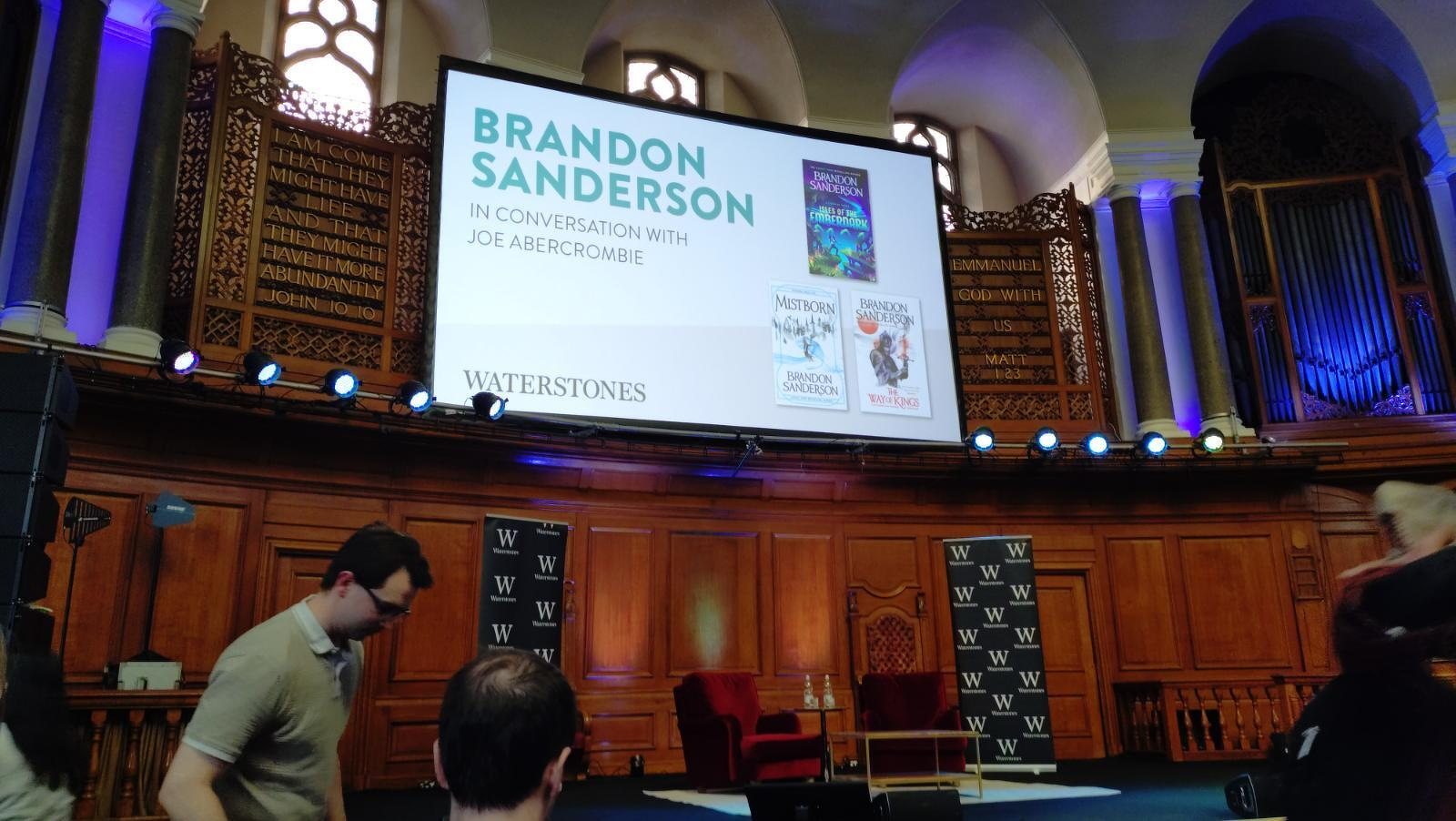 3rd row view, empty stage with two red armchairs,above them is a large screen with the text "Brandon Sanderson in conversation with Joe Abercrombie"