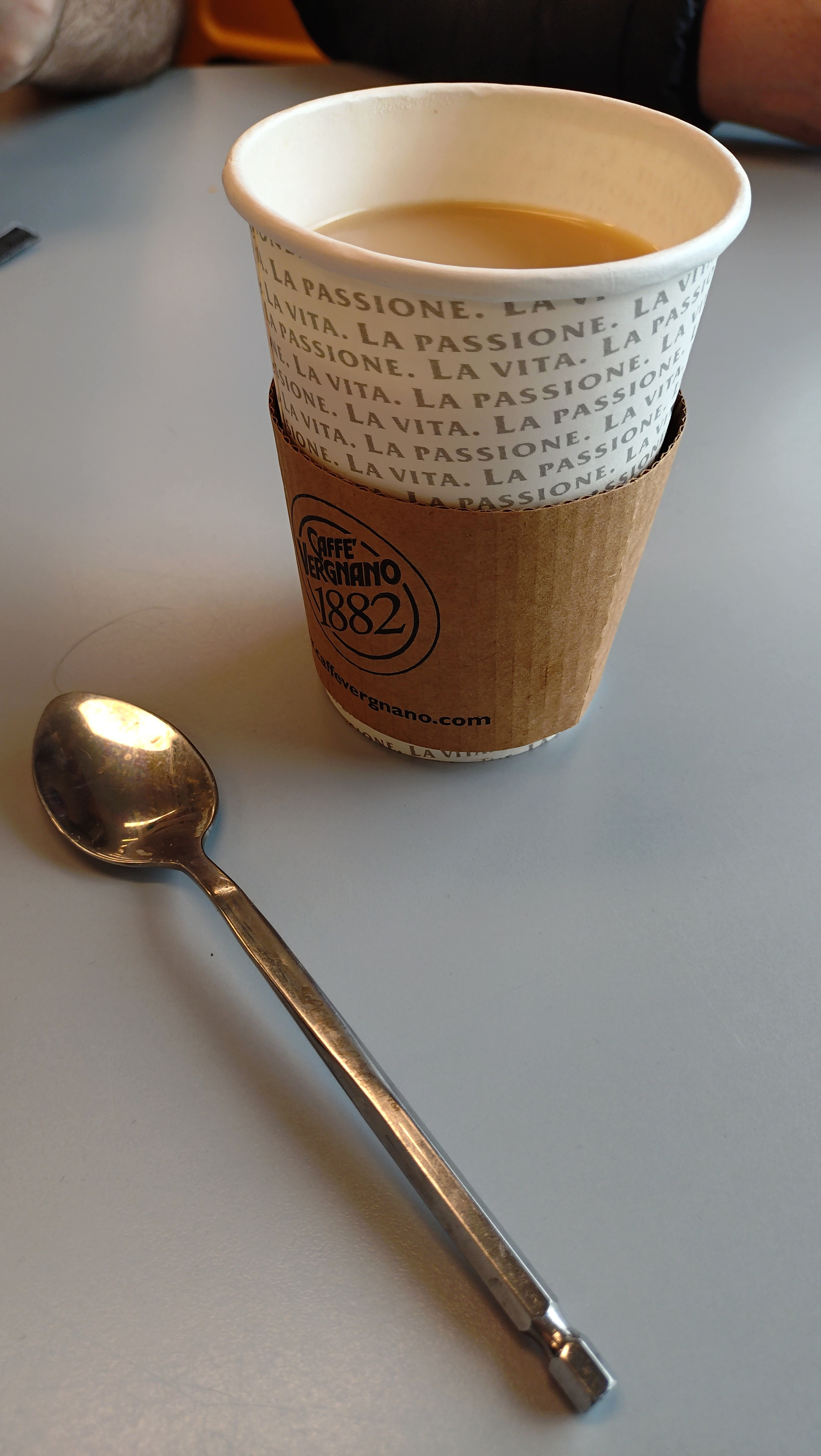 A paper cup filled with hot milky tea (and some sugar) next to a teaspoon with a quarter-inch hex handle (which you could recklessly use in a powered screwdriver if you wanted)