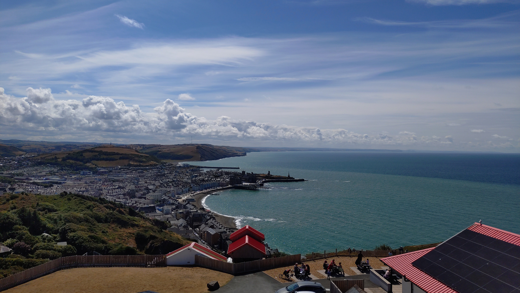 Aberystwyth town spreads out below to the left, the beach, bay and pier are centered, and the sea is on the right. Close up the red roofs of the constitution hill restaurant can be seen, in the far distance fluffy clouds and further coastline under a mostly hazy blue sky&10;