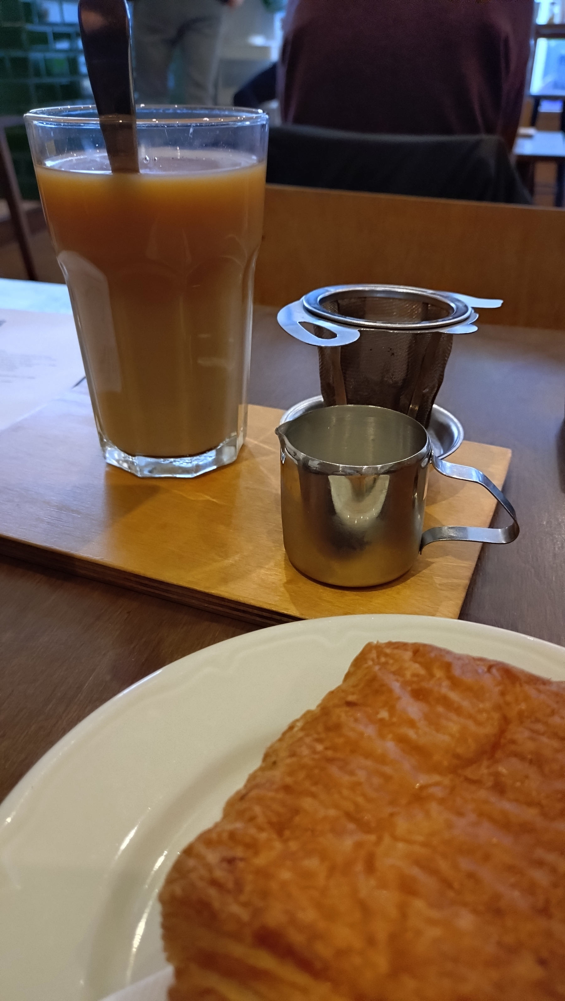 A glass of milky tea with a milk jug, timer and loose tea strainer. A pain au chocolat is in the foreground 